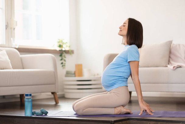 Vue latérale de la jeune femme enceinte faisant backbend pendant la méditation ou l'entraînement de yoga à l'intérieur de la maison. Femme enceinte dans l'étirement occasionnel et faire la pratique de la respiration sur le tapis à l'intérieur - Photo, image
