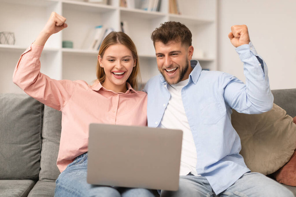 Молодые супруги празднуют победу на ноутбуке в гостиной, мужчина и женщина энергично пожимают кулаки от радости, олицетворяя успех и счастье во время просмотра в Интернете на компьютере дома - Фото, изображение
