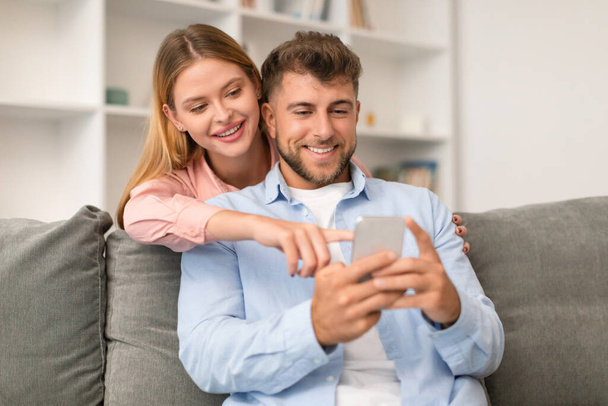Millennial ζευγάρι απορροφάται στο smartphone τους, περιήγηση στα μέσα κοινωνικής δικτύωσης και γραπτών μηνυμάτων, παραδειγματική σύγχρονη επικοινωνία στο άνετο περιβάλλον στο σπίτι, κάθεται στο καθιστικό καναπέ τους - Φωτογραφία, εικόνα
