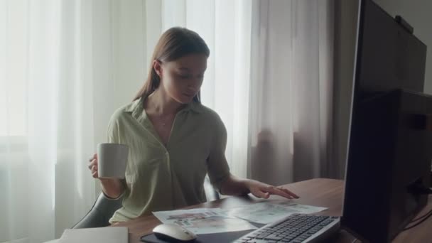 Jonge ondernemer zit aan haar bureau, het hebben van koffie pauze terwijl het kijken door middel van verschillende ontwerpen in de voorkant van haar - Video