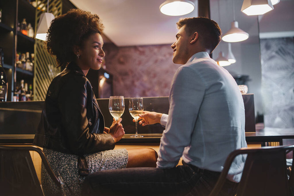 Улыбающаяся межрасовая пара наслаждается романтическим тостом в уютном баре, отмечая особенный момент вместе. - Фото, изображение