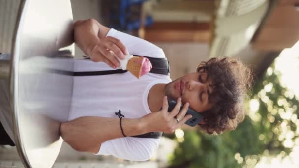 un joven turista árabe con el pelo rizado sostiene un cono de gofre con helado en las manos y lo come y habla por teléfono, se para cerca de un café afuera, los rayos del sol. Vídeo vertical. - Metraje, vídeo