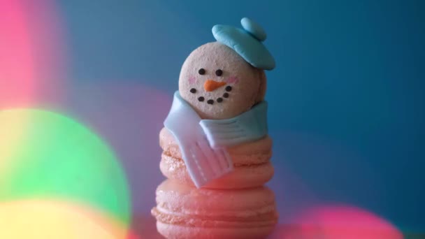 Postre de muñeco de nieve macaron decorado con sombrero y bufanda sobre fondo colorido y bokeh intermitente en primer plano. Feliz Navidad y Feliz Año Nuevo concepto festivo - Metraje, vídeo