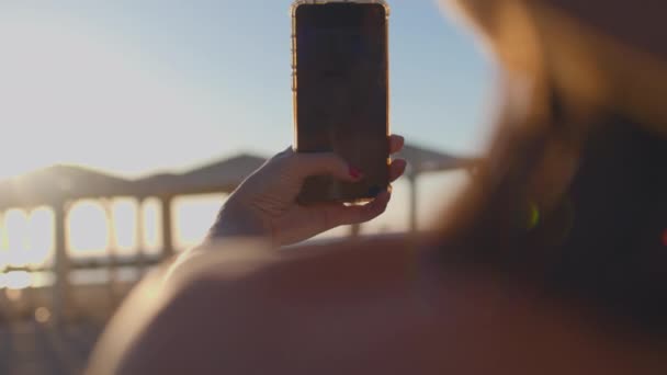 κορίτσι τραβάει βίντεο με ηλιοβασίλεμα στην παραλία. Ένα νεαρό κορίτσι που κάνει σέλβι στις διακοπές. κορίτσι χαλαρώνει στην παραλία. - Πλάνα, βίντεο