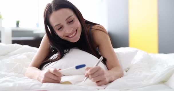 Ευτυχισμένη γυναίκα με τεστ εγκυμοσύνης στην κρεβατοκάμαρα να κρατάει σημειώσεις στο ημερολόγιο. Η πολυαναμενόμενη θετική έννοια της εγκυμοσύνης - Πλάνα, βίντεο