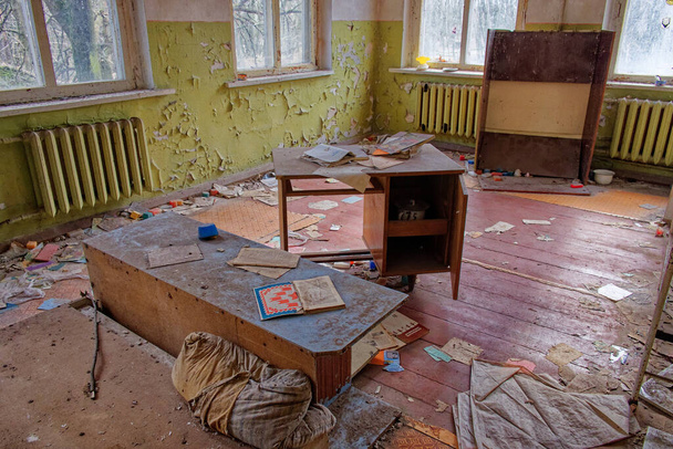 La imagen muestra una habitación abandonada con un escritorio, una estantería y un radiador. Las paredes se pelan y el suelo está cubierto de escombros. Una sala de jardín de infantes abandonada en la zona de exclusión de Chernobyl. - Foto, imagen