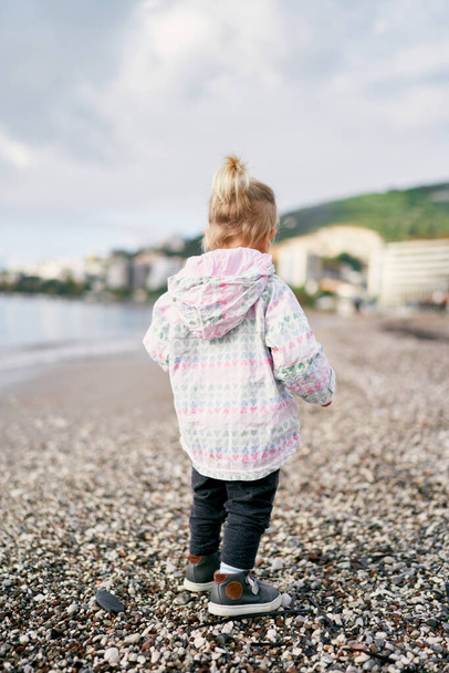 Little girl in a jacket walks along a pebbly beach, looking down at her feet. Back view. High quality photo - Photo, Image