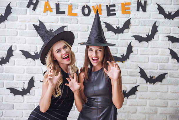 schöne Mädchen in schwarzen Kleidern und Hexenhüten zeigen Krallen, schauen in die Kamera und lächeln, auf einem Hintergrund, der für Halloween dekoriert ist - Foto, Bild
