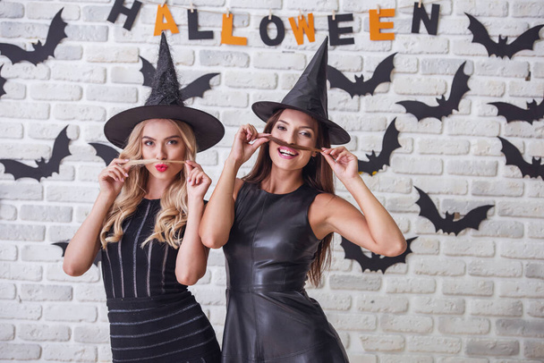 Hermosas chicas en vestidos negros y sombreros de brujas están haciendo bigotes de su cabello, mirando a la cámara y sonriendo, en el fondo decorado para Halloween
 - Foto, imagen