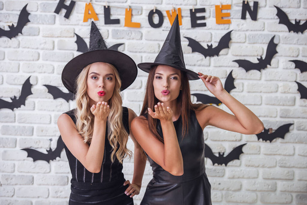 Hermosas chicas en vestidos negros y sombreros de brujas están mirando a la cámara y el envío de besos de aire, en el fondo decorado para Halloween
 - Foto, imagen