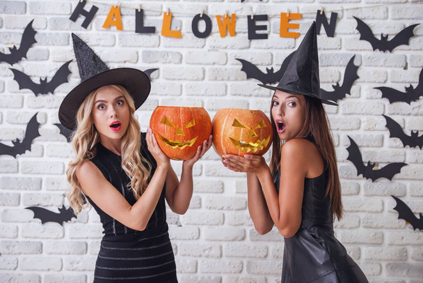 Hermosas chicas en vestidos negros y sombreros de brujas están sosteniendo calabazas de miedo y mirando a la cámara con la boca abierta, en el fondo decorado para Halloween
 - Foto, imagen