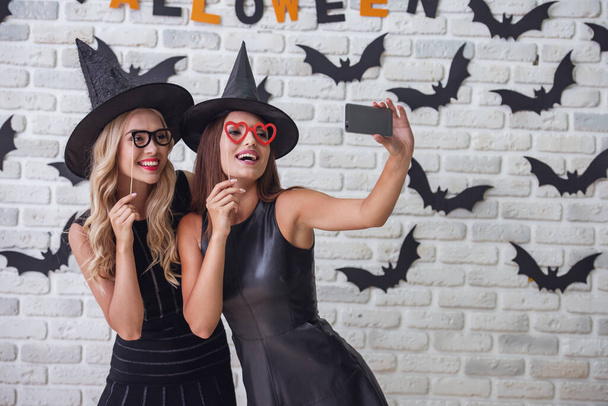 Hermosas chicas en vestidos negros y sombreros de bruja están celebrando accesorios de fiesta y haciendo selfie usando un teléfono inteligente, en el fondo decorado para Halloween
 - Foto, imagen