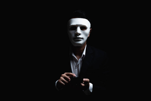 Onbekende zakenman draagt masker met bedekt gezicht met behulp van mobiele telefoon maakt een anonieme oproep intimideren en bedreigen van de gesprekspartner op donkere achtergrond. hacker callcenter concept. - Foto, afbeelding