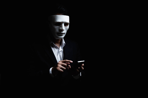 Un homme d'affaires inconnu portant un masque avec un visage couvert à l'aide d'un téléphone portable fait un appel anonyme intimidant et menaçant l'interlocuteur sur fond sombre. concept de centre d'appels pirate. - Photo, image