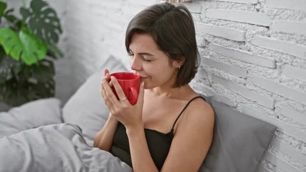 Mujer hispana joven relajada sentada cómodamente en su cama, oliendo el aroma encantador del café de la mañana. hermosa mujer en su dormitorio disfrutando de su bebida con una sonrisa segura. - Metraje, vídeo