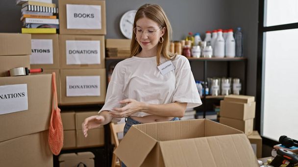 Young blonde woman volunteer packing clothes on cardboard smiling at charity center - Photo, Image