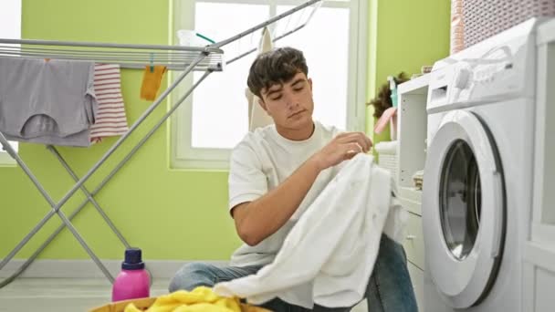 Joven adolescente hispano confiado sonriendo, disfrutando de las tareas domésticas, sosteniendo canasta de ropa sucia y botella de detergente en la lavandería - Metraje, vídeo