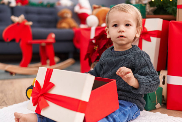 Uroczy blond maluch rozpakowywanie prezent świąteczny siedzi na podłodze w domu - Zdjęcie, obraz