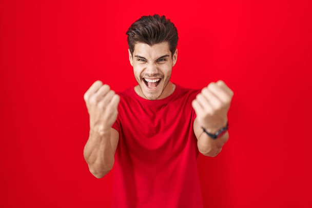 怒りの声を上げながら赤い背景に立っている若いヒスパニック系の男は怒りと怒りの拳を上げる。怒りと攻撃的な考え方.  - 写真・画像