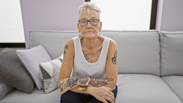 In the cozy comfort of home, a mature, grey-haired woman with glasses, exhibits a serious expression while comfortably sitting and relaxing on her living room sofa, bathed in warm sunlight. - Photo, Image