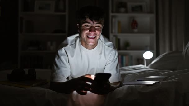 Lächelnder, selbstbewusster junger hispanischer Teenager entspannt sich im Bett, gebadet in weichem Lampenlicht, und tippt mühelos in seinem gemütlichen Schlafzimmer bis spät in die Nacht Nachrichten auf sein Smartphone.. - Filmmaterial, Video