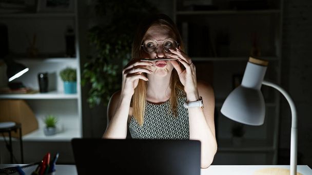 Jeune femme blonde travailleuse d'affaires utilisant un ordinateur portable faisant un geste drôle avec un stylo sur la bouche au bureau - Photo, image