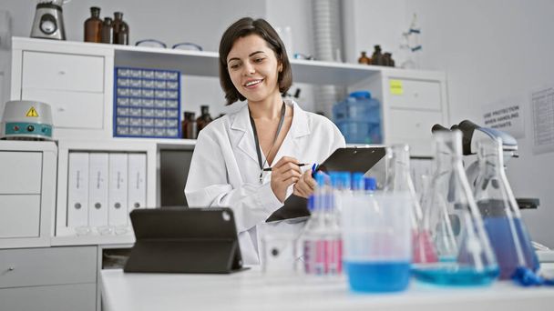 Sonriente joven científico hispano absorto en una videollamada, una hermosa mujer tomando notas en un laboratorio en medio de experimentos - Foto, imagen