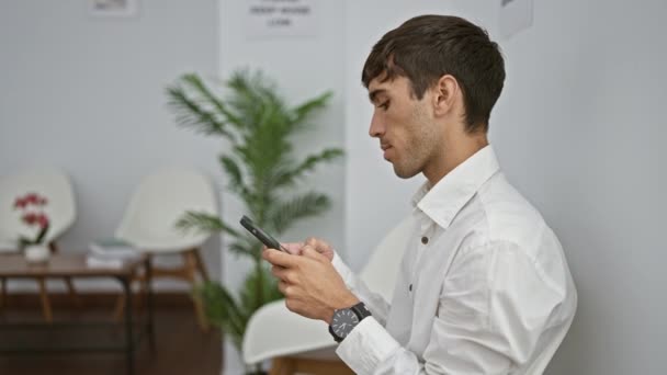 Yakışıklı İspanyol genç adam ciddi ciddi akıllı telefondan iş mesajı yazıyor. Saate bakıyor ve önemli bir toplantı için lobide bekliyor. - Video, Çekim