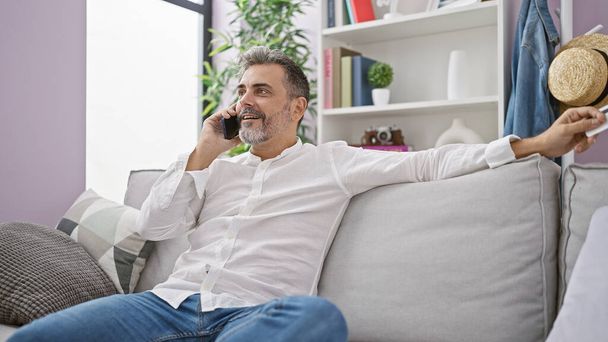 Happy young hispanic man, grey-haired, confidently talks business on smartphone, card in hand, relaxing on home sofa. - Photo, Image