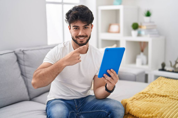 Homem hispânico com barba usando touchpad sentado no sofá alegre com um sorriso no rosto apontando com a mão e o dedo para o lado com expressão feliz e natural  - Foto, Imagem