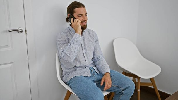 Молодой латиноамериканец разговаривает на смартфоне, сидя на стуле и улыбаясь в зале ожидания. - Фото, изображение