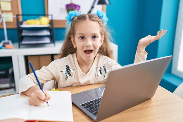 Niña sentada sobre la mesa haciendo deberes con laptop celebrando el logro con sonrisa feliz y expresión ganadora con la mano levantada  - Foto, imagen