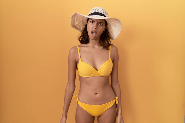 Mulher hispânica jovem usando biquíni e chapéu de verão em face de choque, parecendo cético e sarcástico, surpreso com a boca aberta  - Foto, Imagem