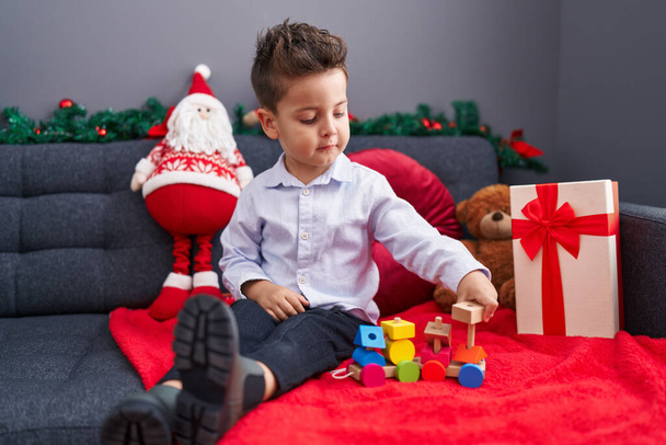 Uroczy latynoski maluch bawiący się zabawką z pociągu siedzący na kanapie przez świąteczną dekorację w domu - Zdjęcie, obraz