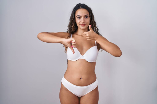 Jonge Spaanse vrouw met witte lingerie die duimen op en neer doet, onenigheid en eensgezindheid. gek conflict  - Foto, afbeelding