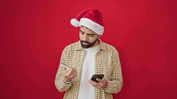 孤立した赤い背景の上にクリスマスの帽子をかぶっているスマートフォンおよびクレジット カードを使用して若いヒスパニックの男 - 写真・画像