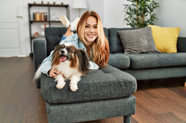 Νεαρή καυκάσια γυναίκα χαμογελά αυτοπεποίθηση που βρίσκεται στον καναπέ με το σκυλί στο σπίτι - Φωτογραφία, εικόνα