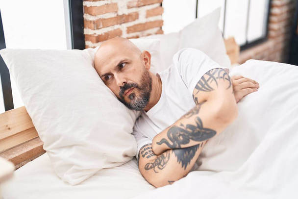Young bald man stressed sitting on bed at bedroom - 写真・画像
