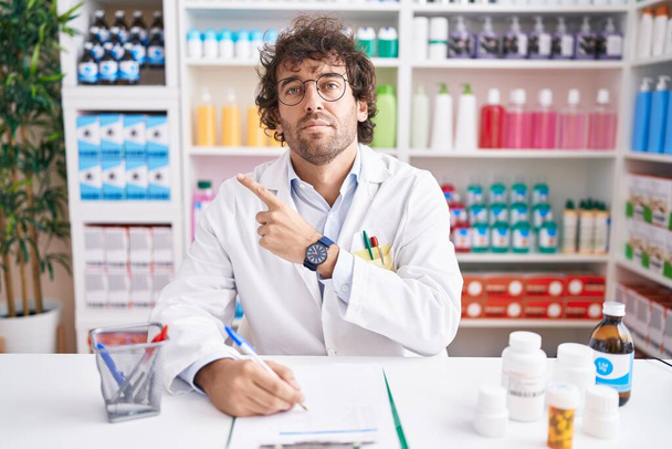 Hispán fiatalember dolgozik gyógyszertár gyógyszertár gyógyszertár mutató kéz ujját oldalra mutató reklám, komoly és nyugodt arc  - Fotó, kép