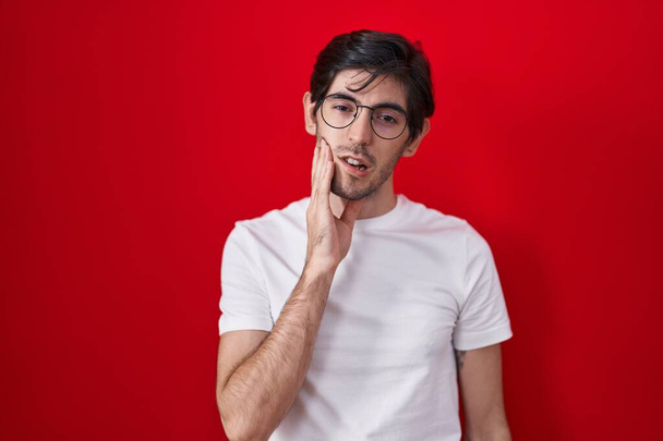 Homem hispânico jovem em pé sobre fundo vermelho tocando a boca com a mão com expressão dolorosa por causa da dor de dente ou doença dentária nos dentes. dentista  - Foto, Imagem