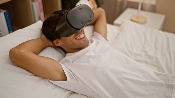 Feliz chico hispano joven cautivado por el juego en línea, disfrutando de auriculares vr futuristas, acostado en el dormitorio con gadget, confiado en su capacidad de juego, entusiasta de interior con sonrisa radiante. - Foto, imagen