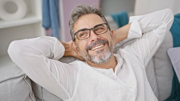 At home, a relaxed young hispanic man with grey hair is joyfully sitting on the sofa, hands on head conveying comfort and confidence - Photo, Image