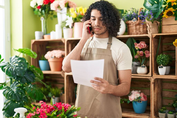 Молодий латинський чоловік-флорист розмовляє на смартфоні читаючи документ у квітковому магазині
 - Фото, зображення