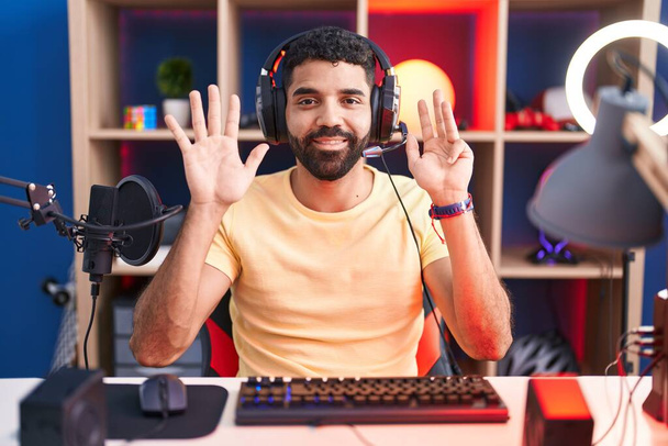 Latynos z brodą grający w gry wideo ze słuchawkami pokazującymi i wskazującymi palcami numer dziewięć uśmiechając się z uśmiechem pewny siebie i szczęśliwy.  - Zdjęcie, obraz