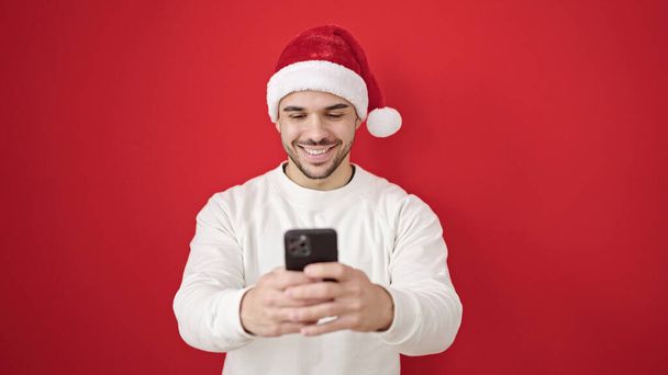 孤立した赤い背景の上にスマートフォンを使用してクリスマスの帽子をかぶっている若いヒスパニック男性 - 写真・画像