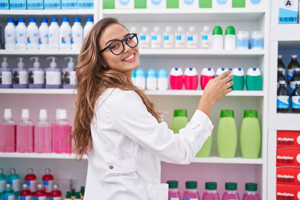 Giovane bella donna ispanica farmacista sorridente fiducioso bottiglia tenuta sugli scaffali in farmacia - Foto, immagini