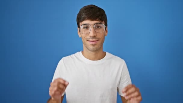 Молодий іспаномовний чоловік бере окуляри посміхаючись впевнено над ізольованим синім тлом - Кадри, відео