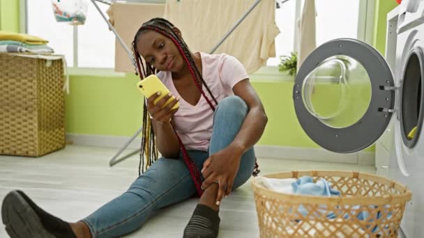 Linda mulher afro-americana sentada no chão da lavanderia, sorrindo enquanto ela mensagens em seu smartphone em meio a tarefas domésticas - Filmagem, Vídeo
