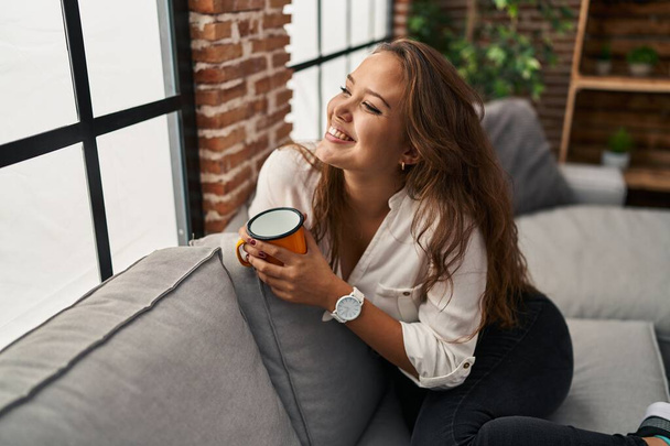 Νεαρή όμορφη ισπανίδα γυναίκα πίνει καφέ καθισμένη στον καναπέ στο σπίτι - Φωτογραφία, εικόνα