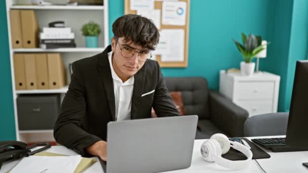 Уверенный молодой латиноамериканец пожимает руку рассеянной позитивности на работе, стоя, ноутбук под рукой в оживленной офисной среде - Кадры, видео